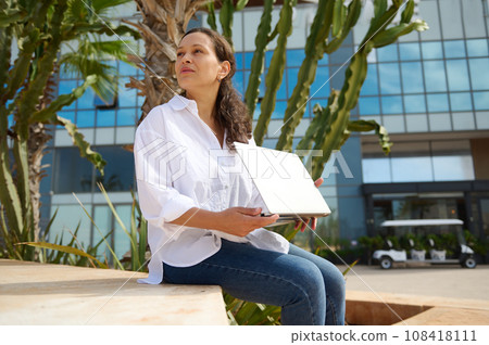 Confident young Latina woman in white shirt and blue casual jeans, holding laptop and looking into the distance 108418111