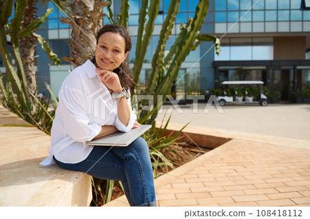 Beautiful woman in casual clothes, sitting on a stone bench with laptop on her knees, smiling cutely looking at camera 108418112
