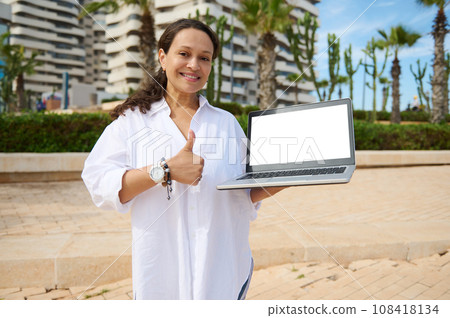 Smiling sales manager showing laptop mockup screen, thumb up against buildings background with developed infrastructure Smiling sales manager showing laptop mockup screen, thumb up against buildings background with developed infrastructure 108418134