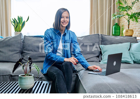 Young woman sitting on living room sofa at home with laptop Young woman sitting on living room sofa at home with laptop 108418613
