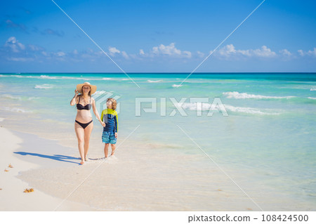 A radiant pregnant mother and her excited son share a tender moment on a serene, snow-white beach, celebrating family love amidst nature's beauty 108424500