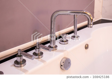 Closeup of silver faucet for Hot water Cold water on a white bathtub with circular windows and trees in the background. 108425186
