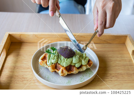 Hands holding hands and a fork prepare to cut green tea waffles in a white plate on a traditional Japanese wooden tray to eat. Hands holding hands and a fork prepare to cut green tea waffles in a white plate on a traditional Japanese wooden tray to eat. 108425225