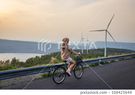 Woman wearing cute outfit riding bicycle at sunset with wind turbine mountains and rivers in the background 108425649