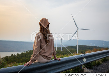 Woman sitting looking at sunset with mountains and river and wind turbine in the background 108425651