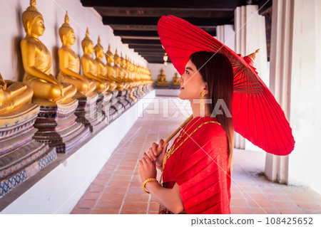 Young woman wearing traditional red Thai dress and golden accessories stands holding a traditional umbrella in the historical site Wat Phutthaisawan Ayutthaya. Thai national costume 108425652