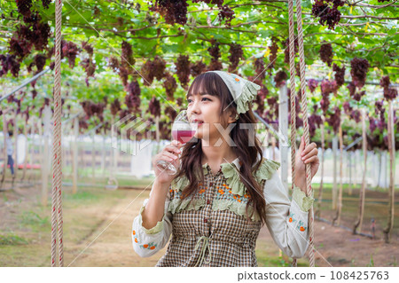 Woman wearing cute dress stand drinking grape juice or wine in vineyard smiling happily Woman wearing cute dress stand drinking grape juice or wine in vineyard smiling happily 108425763