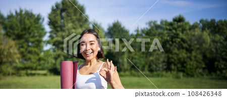 Excited young woman standing with sports mat, yoga clothes, shows okay sign, workout in park, wellbeing training session outdoors 108426348