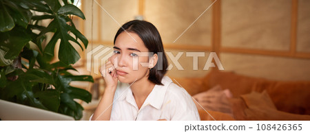 Vertical shot of sad and gloomy young woman, sitting with laptop in cafe, grimacing and looking upset, feeling tired or disappointed Vertical shot of sad and gloomy young woman, sitting with laptop in cafe, grimacing and looking upset, feeling tired or disappointed 108426365