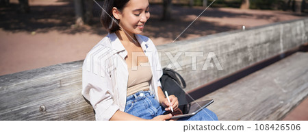 Young asian creative girl drawing with pen on graphic tablet, sitting outdoors in park, painting smth 108426506