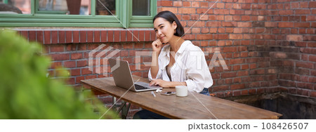 Beautiful brunette woman sitting outdoors and using laptop, chatting online, working with computer outside office Beautiful brunette woman sitting outdoors and using laptop, chatting online, working with computer outside office 108426507