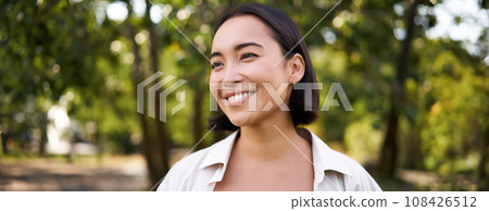 People concept. Smiling asian girl looking at camera with happy, joyful emotion, posing in green park on a warm summer day People concept. Smiling asian girl looking at camera with happy, joyful emotion, posing in green park on a warm summer day 108426512