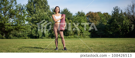 Confident and sporty young asian woman, standing in park with rubber mat, wearing sportswoman, smiling at camera, workout on fresh air 108426513