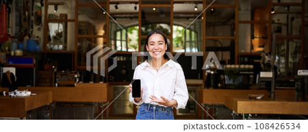 Enthusiastic young asian woman, cafe manager demonstrating app on mobile phone, showing smartphone screen, standing in front of cafe business 108426534