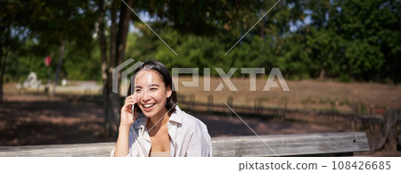 Portrait of happy young asian woman talking on mobile phone in park, sitting on bench and having a telephone call, chatting lively 108426685