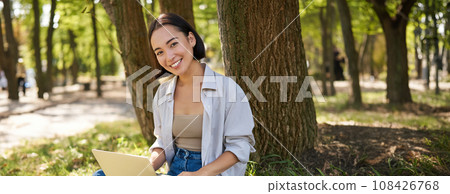 Beautiful asian girl sitting in park with laptop, working on remote, typing on keyboard, smiling at camera, resting beside tree 108426768