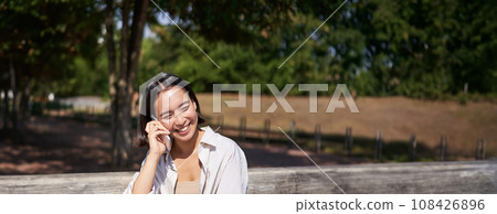 Smiling asian girl relaxing in park, talking on smartphone, having a mobile call while resting outdoors Smiling asian girl relaxing in park, talking on smartphone, having a mobile call while resting outdoors 108426896