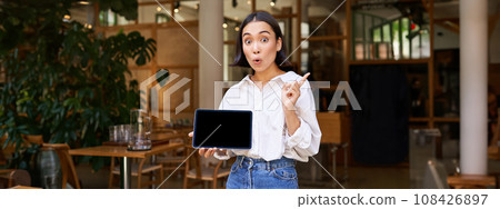 Smiling asian businesswoman, demonstrating, showing tablet screen and pointing at banner, advertising Smiling asian businesswoman, demonstrating, showing tablet screen and pointing at banner, advertising 108426897