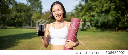 Smiling asian fitness girl with rubber yoga mat, shows her smartphone screen, recommends workout application, stands on lawn in park 108426902