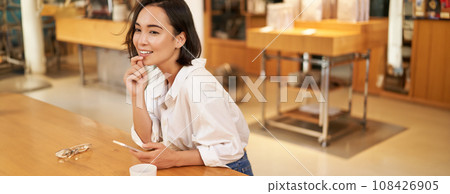 Portrait of beautiful asian woman with smartphone, relaxing in cafe, sitting and enjoying coffee while using mobile phone Portrait of beautiful asian woman with smartphone, relaxing in cafe, sitting and enjoying coffee while using mobile phone 108426905