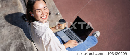 Beautiful young asian female student, sitting with laptop on bench in park, drinking takeaway coffee and listening music, working remotely, studying outdoors 108426909