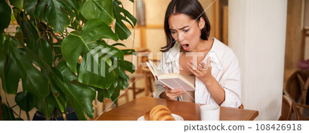 Portrait of asian woman looking shocked at book pages, reading something interesting, concentrating, sitting in cafe with coffee and croissant 108426918