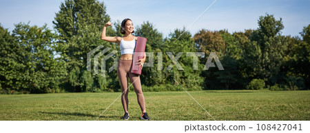 Portrait of fit and healthy young fitness woman, shows her biceps with pleased smiling face, stands with yoga rubber mat on grass in park Portrait of fit and healthy young fitness woman, shows her biceps with pleased smiling face, stands with yoga rubber mat on grass in park 108427041