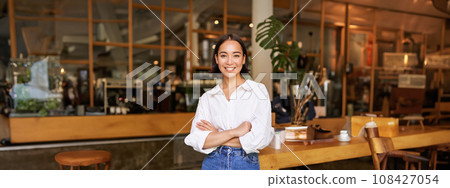 Portrait of young female entrepreneur, cafe owner, standing at the door entrance and smiling, looking confident 108427054