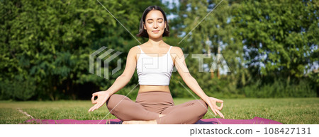 Sporty woman meditating on fresh air, sitting on fitness mat and practice yoga, smiling pleased. Sport and people concept 108427131