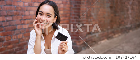 Beautiful young asian woman with credit card, sitting near laptop and smiling, paying bills, shops online, order smth on computer 108427164