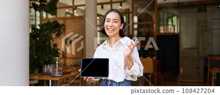 Friendly smiling asian woman, cafe manager, showing tablet screen and looking happy, demonstrating her restaurant profit 108427204