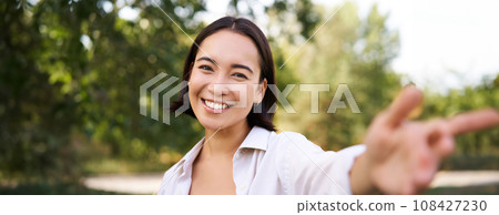 People and lifestyle. Happy asian woman takes selfie in park, photo on smartphone, smiling and looking joyful People and lifestyle. Happy asian woman takes selfie in park, photo on smartphone, smiling and looking joyful 108427230