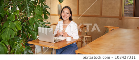 Vertical shot of young asian girl sits in cafe with laptop and smartphone, relaxing and surfing the net, working remotely Vertical shot of young asian girl sits in cafe with laptop and smartphone, relaxing and surfing the net, working remotely 108427311