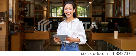 Smiling beautiful waitress, businesswoman with digital tablet, laughing and looking happy, standing in front of cafe or restaurant Smiling beautiful waitress, businesswoman with digital tablet, laughing and looking happy, standing in front of cafe or restaurant 108427373