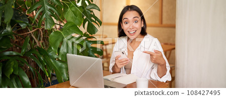 Amazed asian girl points at her phone and looks surprised, tells big news, sits in cafe with laptop 108427495