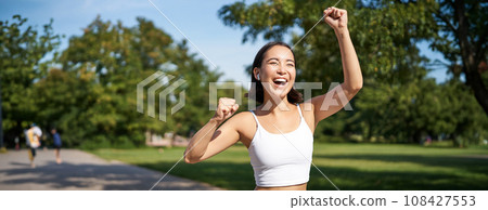 Happy fitness girl achieve goal, finish marathon, running with hands up, celebrating victory while jogging, triumphing in park Happy fitness girl achieve goal, finish marathon, running with hands up, celebrating victory while jogging, triumphing in park 108427553