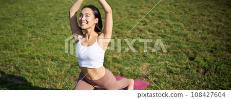Portrait of asian young woman smiling pleased, stretching her body in yoga asana, standing on mat and workout in park Portrait of asian young woman smiling pleased, stretching her body in yoga asana, standing on mat and workout in park 108427604