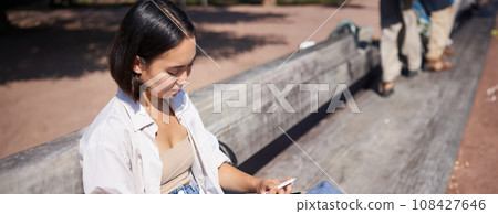 Young asian creative girl drawing with pen on graphic tablet, sitting outdoors in park, painting smth 108427646