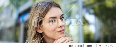 Close up portrait of young woman, tuck hair behind ear, looking flirty and smiling, sitting in blue shirt outdoors on street 108427770