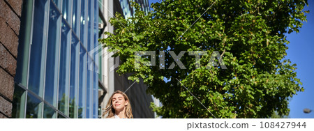 Portrait of confident saleswoman in her 30s, posing near office building on street, wearing beige suit, holding folder with documents Portrait of confident saleswoman in her 30s, posing near office building on street, wearing beige suit, holding folder with documents 108427944