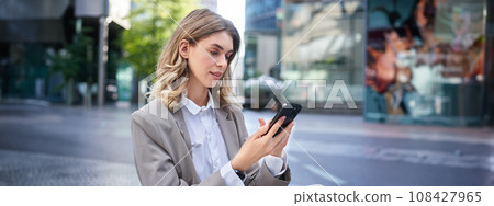 Blond woman in suit, looking at her mobile phone, order taxi, using application, posing outdoors 108427965