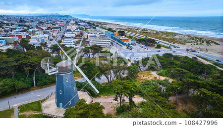 Aerial blue murphy windmill with white blades overlooking city of hoses and ocean with beach 108427996