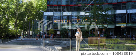 Corporate businesswoman in beige suit. Silhouette of young corporate woman with documents and laptop, posing outdoors in city center Corporate businesswoman in beige suit. Silhouette of young corporate woman with documents and laptop, posing outdoors in city center 108428019