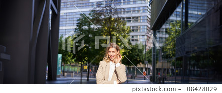 Portrait of smiling businesswoman in corporate clothing, looking confident and happy, wearing beige suit, standing outdoors on street, outside office 108428029