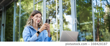 Portrait of saleswoman working outside office, sititng with laptop, using mobile phone. Digital nomad woman studying, using computer outdoors 108428187