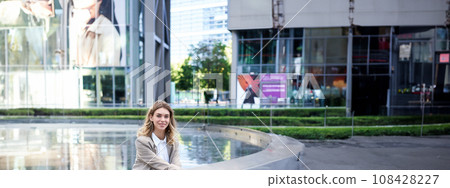 Vertical shot of successful businesswoman in suit, sitting near fountain and business hall, smiling at camera with confidence Vertical shot of successful businesswoman in suit, sitting near fountain and business hall, smiling at camera with confidence 108428227