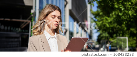 Portrait of corporate woman reads news, works on her digital tablet while on her way to office, stansd on street of city center in suit Portrait of corporate woman reads news, works on her digital tablet while on her way to office, stansd on street of city center in suit 108428258