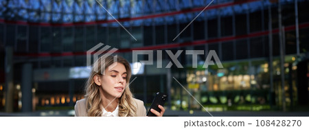 Vertical shot of businesswoman drinks coffee, looks at mobile phone app. Corporate woman on her lunch break, using smartphone 108428270