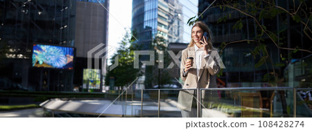 Corporate woman in beige suit, drinks morning coffee, looks at her smartphone screen, uses application, checking messages on phone, stands on street outside office 108428274
