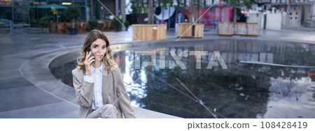 Vertical shot of corporate woman sitting outdoors with laptop, talking on mobile phone, working ouside office building while waiting for someone 108428419